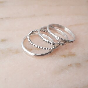 May include: A set of silver rings in different styles. One ring is plain, another has small beads, a third has an etched design, and the last has a hammered finish. The rings are arranged on a pale pink surface.