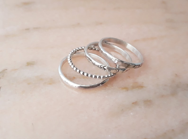 May include: A set of silver rings in different styles. One ring is plain, another has small beads, a third has an etched design, and the last has a hammered finish. The rings are arranged on a pale pink surface.