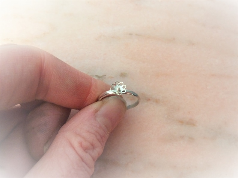 May include: A silver ring featuring a flower design. The ring is held in a hand, highlighting the floral detail. The ring's band is slender and polished, and the flower has multiple petals. The ring is a piece of jewellery.