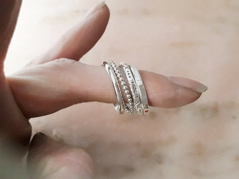 May include: A selection of silver rings stacked on a finger. The rings feature diverse designs, including a beaded band, a smooth band, and bands with etched patterns. The rings are set against a soft, neutral backdrop.