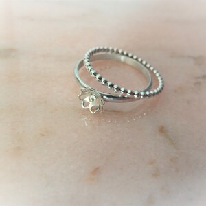 May include: Two silver rings. One ring has a floral design, the other a beaded texture. The rings are on a light pink marble surface, creating a soft and elegant aesthetic. The rings are likely made of silver.
