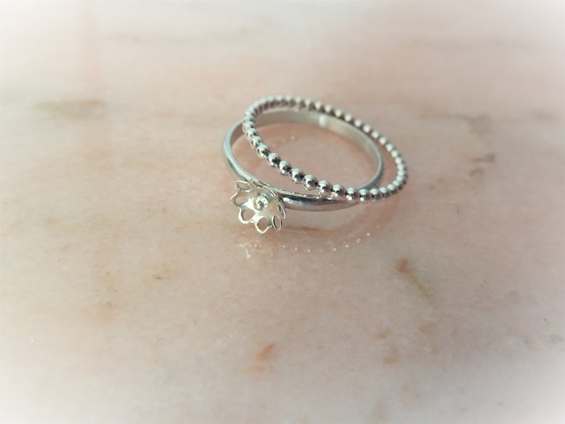 May include: Two silver rings. One ring has a floral design, the other a beaded texture. The rings are on a light pink marble surface, creating a soft and elegant aesthetic. The rings are likely made of silver.