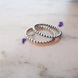 May include: Three silver rings with a beaded design. The rings are stacked, creating a layered effect. The rings are made of a shiny metal and are set against a light pink and white background.