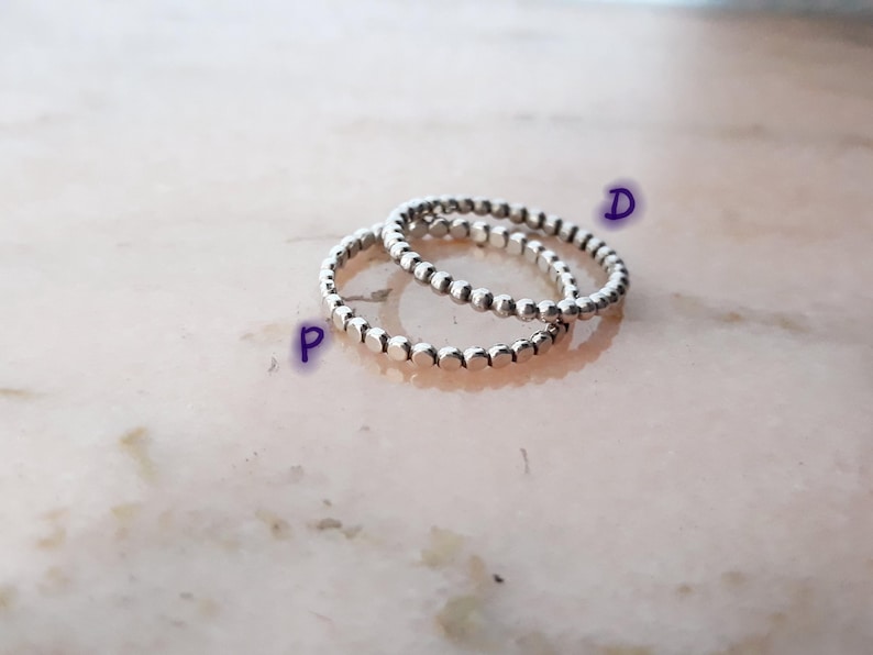 May include: Three silver rings with a beaded design. The rings are stacked, creating a layered effect. The rings are made of a shiny metal and are set against a light pink and white background.