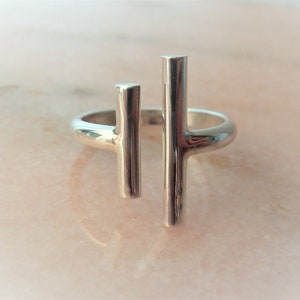 Op de afbeelding: Een zilveren ring met twee verticale staven, waarvan er één iets korter is dan de andere, wat een minimalistisch ontwerp creëert.