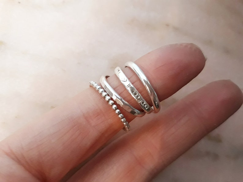 May include: A selection of silver rings on a finger. The rings include a beaded band, a hammered band, and several smooth bands with etched designs. The rings are made of a shiny, metallic material.