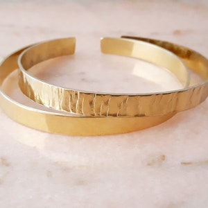Peut inclure: Deux bracelets en métal doré, l'un avec une finition lisse et l'autre avec une texture martelée.
