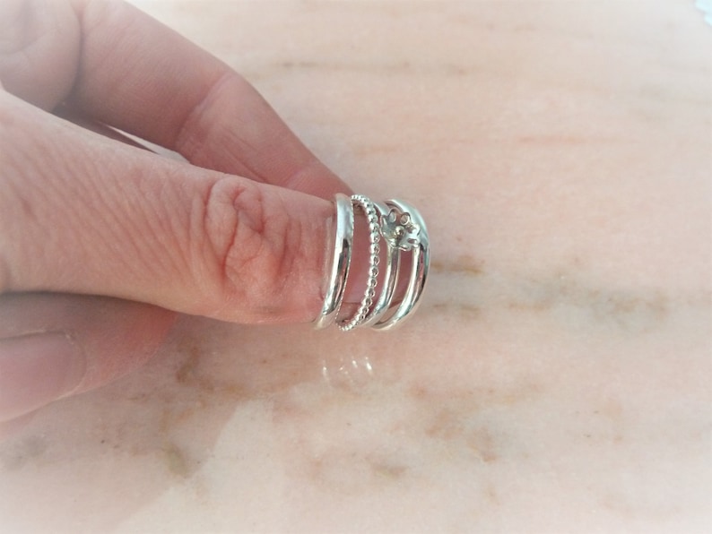 May include: A set of four silver rings. One ring is a solid band, another has a beaded design, and the third features a floral design. The rings are worn together on a finger.