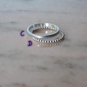 May include: Two silver rings are stacked. The top ring is smooth, the bottom ring has a beaded design. The rings reflect light, set against a blurred, light-coloured background. The rings are likely made of sterling silver.