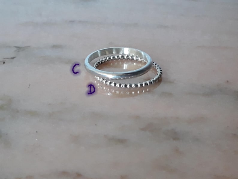 May include: Two silver rings are stacked. The top ring is smooth, the bottom ring has a beaded design. The rings reflect light, set against a blurred, light-coloured background. The rings are likely made of sterling silver.