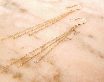 May include: A pair of gold-toned dangle earrings featuring three delicate, thin chain strands suspended from a curved ear wire hook. The earrings have a minimalist design with multiple fine metal links that hang freely to create a layered effect.