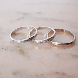 May include: Three silver rings are shown. The rings feature varied designs: one with a faceted texture, another with etched geometric patterns, and a third with a smooth, polished surface. They are set against a light pink background.