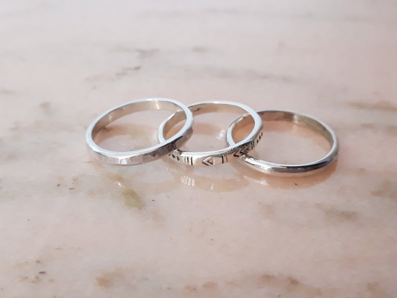 May include: Three silver rings are shown. The rings feature varied designs: one with a faceted texture, another with etched geometric patterns, and a third with a smooth, polished surface. They are set against a light pink background.