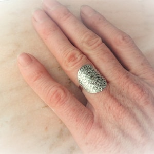 Könnte beinhalten: Ein silberner Ring mit einem großen, ovalen, detaillierten Design. Der Ring wird an einem Finger getragen.