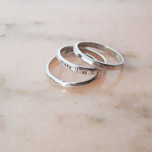 May include: Three silver rings are stacked on a light pink surface. One ring has a hammered texture, another has a geometric pattern, and the third has a smooth, polished finish. The rings are likely for everyday wear.