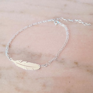 Peut inclure: Un bracelet en argent délicat avec un charm en forme de plume. Le bracelet a un design à double chaîne et un fermoir réglable. Le charm en forme de plume est de couleur or clair et est le point central. Le bracelet est présenté sur une surface rose clair.