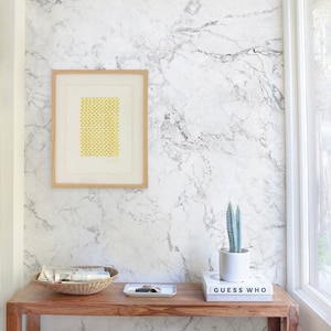 May include: A wooden console table with a white marble wallpaper backdrop. The table has a framed print with a yellow geometric pattern, a wicker basket with magazines, a small silver tray, a potted cactus, and two books titled "Guess Who" and "Interiors: Modern & Classic".