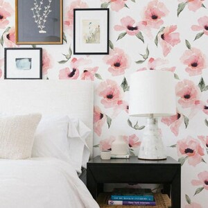 May include: A white bedroom with a bed, a nightstand, and a lamp. The wall is covered in pink floral wallpaper. The bed has a white duvet and a white pillow. The nightstand is black and has a book on top of it. The lamp is white and has a white shade.