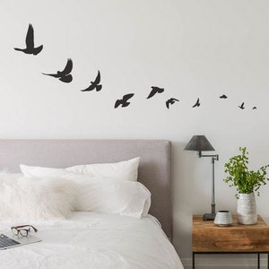 May include: A wall decal featuring a flock of nine black birds in flight, arranged in a diagonal formation. The silhouettes vary in size, with the largest bird on the far left and progressively smaller birds trailing toward the right side.