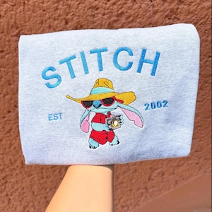 May include: A light gray sweatshirt featuring the word "STITCH" in blue, with an illustration of Stitch wearing sunglasses and a hat. The text "EST 2002" is also visible. The sweatshirt is held up by a person.