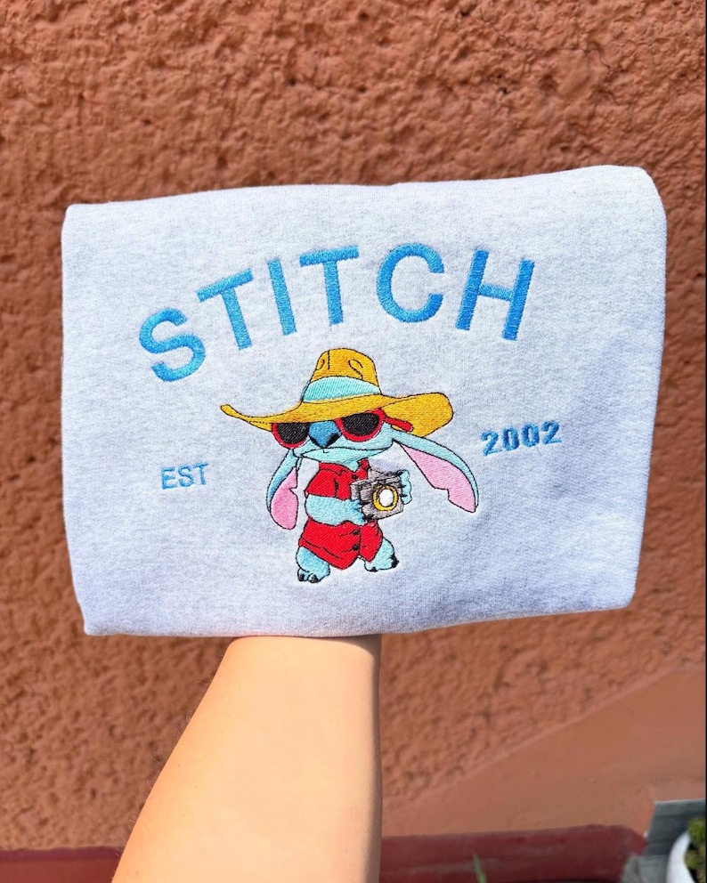 May include: A light gray sweatshirt featuring the word "STITCH" in blue, with an illustration of Stitch wearing sunglasses and a hat. The text "EST 2002" is also visible. The sweatshirt is held up by a person.