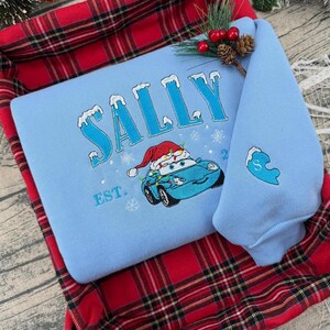 Puede incluir: Sudadera azul claro con el nombre "SALLY" bordado en azul con detalles de nieve. Presenta un coche de dibujos animados con un gorro de Pap&aacute; Noel y luces navide&ntilde;as. La manga tiene un coraz&oacute;n azul con la letra "S".