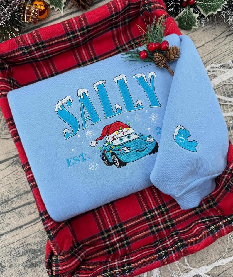 Puede incluir: Sudadera azul claro con el nombre "SALLY" bordado en azul con detalles de nieve. Presenta un coche de dibujos animados con un gorro de Pap&aacute; Noel y luces navide&ntilde;as. La manga tiene un coraz&oacute;n azul con la letra "S".