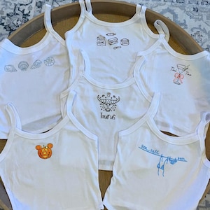 Puede incluir: Camisetas sin mangas blancas con varios diseños gráficos en negro y naranja. Los diseños incluyen conchas marinas, ingredientes para hornear, un símbolo del zodiaco Tauro, un cóctel, una calabaza y el texto "Sea, salt, sun."