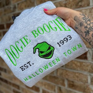 K&ouml;nnte beinhalten: Ein hellgraues Sweatshirt mit den gr&uuml;nen Worten "OOGIE BOOGIE", einem gr&uuml;nen Gesichts-Design und dem Text "1993 EST. HALLOWEEN TOWN". Das Sweatshirt ist gefaltet und wird gehalten.