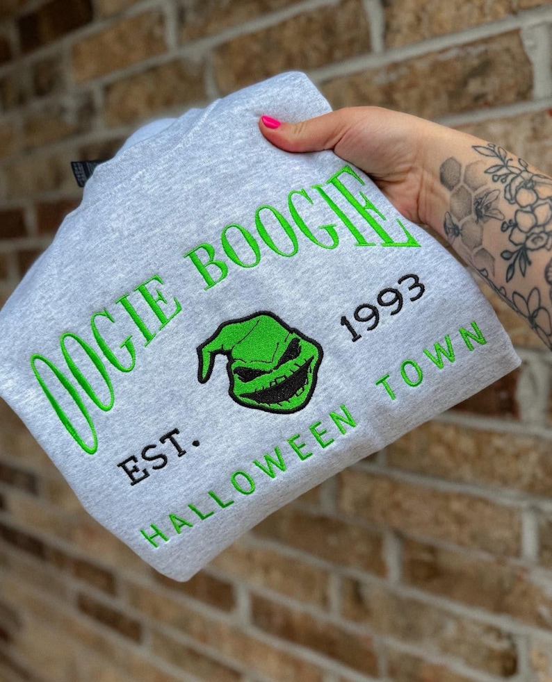 K&ouml;nnte beinhalten: Ein hellgraues Sweatshirt mit den gr&uuml;nen Worten "OOGIE BOOGIE", einem gr&uuml;nen Gesichts-Design und dem Text "1993 EST. HALLOWEEN TOWN". Das Sweatshirt ist gefaltet und wird gehalten.