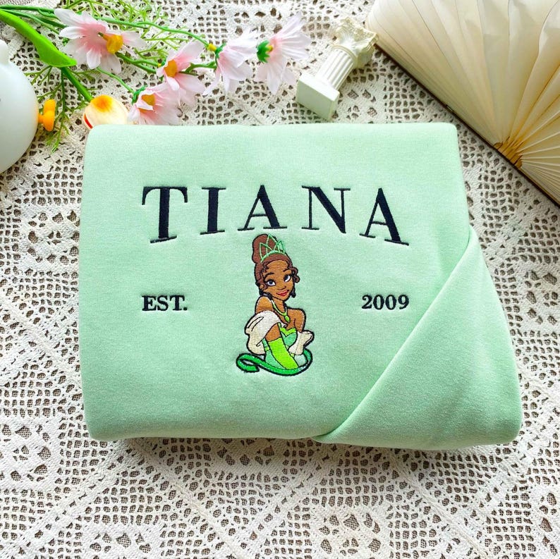 Może przedstawiać: Jasnozielona bluza z haftowanym czarnym napisem "TIANA". Poniżej haftowany wizerunek księżniczki, z napisem "EST. 2009" po obu stronach. Bluza jest złożona, ukazując swoją miękką fakturę.