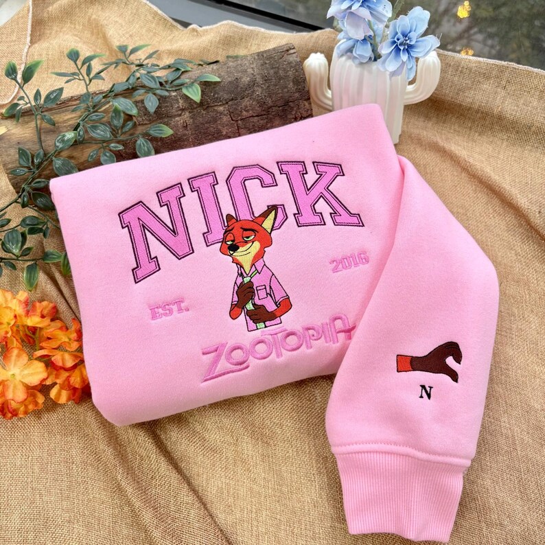May include: A pink sweatshirt featuring the word "NICK" in large, pink, arched lettering, with an illustration of a fox in a pink shirt. The word "ZOOTOPIA" is embroidered below the fox. The sleeve has a brown paw print and the letter "N".