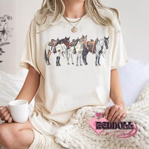 May include: A white t-shirt with a graphic of four horses wearing saddles. The horses are brown, white, and black. The t-shirt has a crew neck and short sleeves.