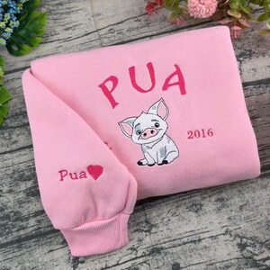 Pode incluir: Uma camisola rosa claro com texto bordado em rosa escuro que diz PUA acima de uma ilustra&ccedil;&atilde;o de um porquinho. O ano 2016 est&aacute; bordado &agrave; direita do porquinho. A manga apresenta o texto Pua com um pequeno &iacute;cone de cora&ccedil;&atilde;o rosa escuro.