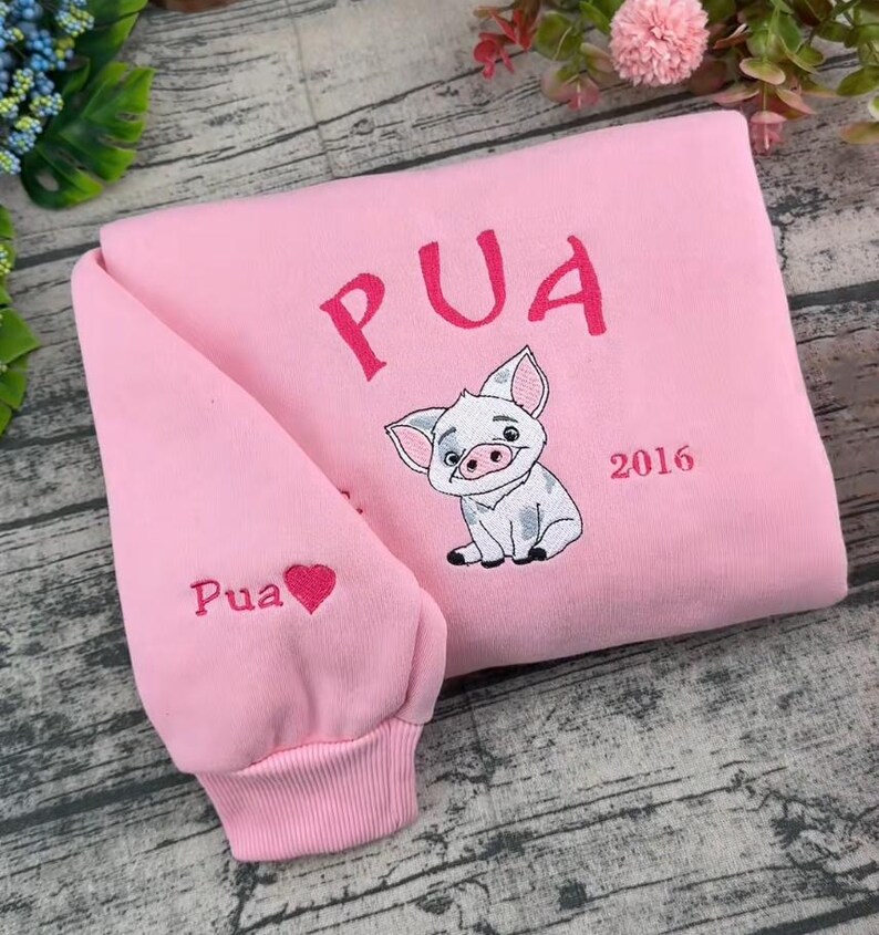 Pode incluir: Uma camisola rosa claro com texto bordado em rosa escuro que diz PUA acima de uma ilustra&ccedil;&atilde;o de um porquinho. O ano 2016 est&aacute; bordado &agrave; direita do porquinho. A manga apresenta o texto Pua com um pequeno &iacute;cone de cora&ccedil;&atilde;o rosa escuro.