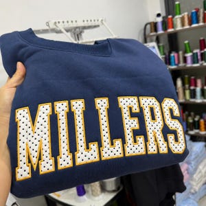 Può includere: Una felpa girocollo blu navy con la parola MILLERS ricamata in grandi lettere stampatello. Le lettere sono bianche con un motivo a pois neri e sono delineate da un filo spesso giallo oro, creando un aspetto audace e strutturato.