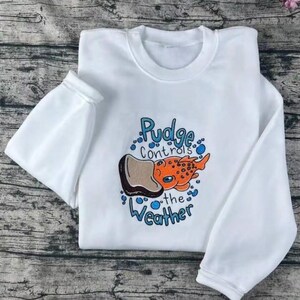 K&ouml;nnte beinhalten: Wei&szlig;es Sweatshirt mit Rundhalsausschnitt und langen &Auml;rmeln. Auf der Vorderseite befindet sich eine Cartoon-Illustration eines orangefarbenen und braunen Fisches mit dem Text "Pudge controls the Weather" in Blau.