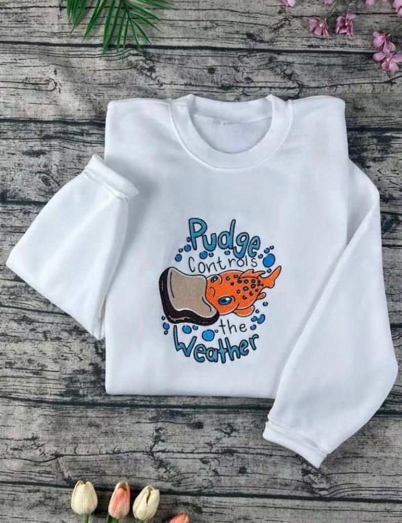 K&ouml;nnte beinhalten: Wei&szlig;es Sweatshirt mit Rundhalsausschnitt und langen &Auml;rmeln. Auf der Vorderseite befindet sich eine Cartoon-Illustration eines orangefarbenen und braunen Fisches mit dem Text "Pudge controls the Weather" in Blau.