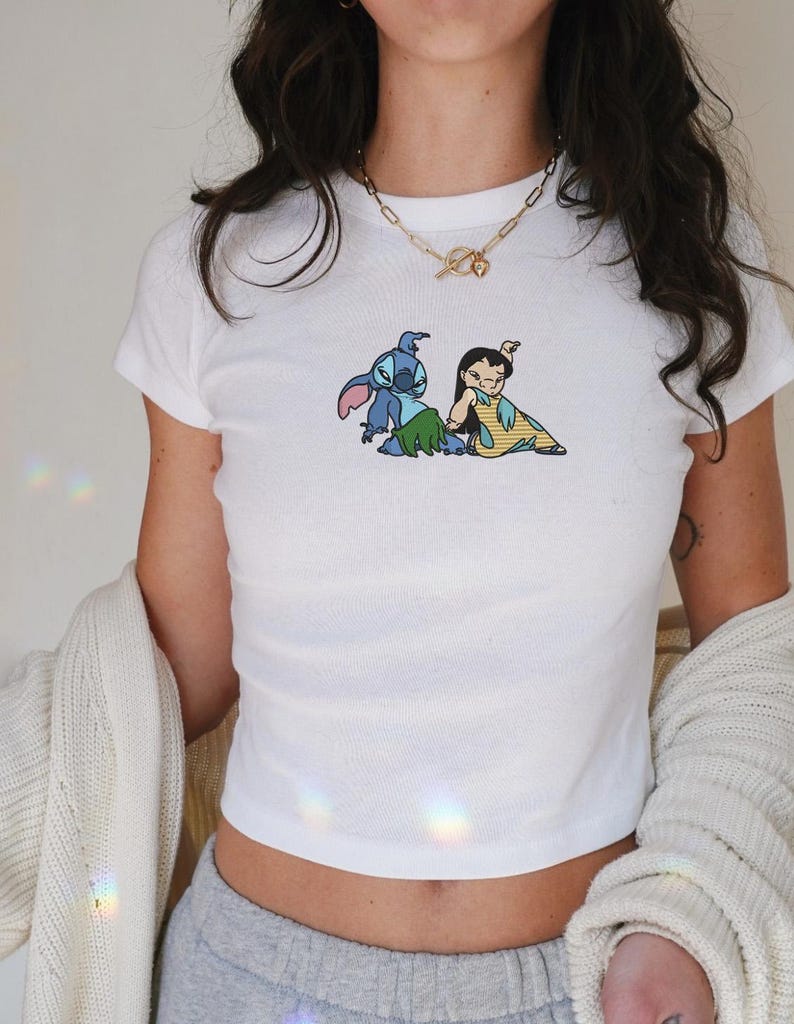 May include: White, short-sleeved crop top with an embroidered design of Stitch and Lilo from Lilo & Stitch. The design shows Stitch with a grass skirt and Lilo in a yellow dress. The top is ribbed and form-fitting. The top is a popular item.