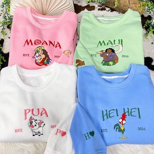 May include: Four crewneck sweatshirts in pink, light green, white, and blue. Each features embroidered character art and text: Moana, Maui, Pua, or Hei Hei. The text includes names, EST. 2016, and small sleeve details like a heart or hand icon.