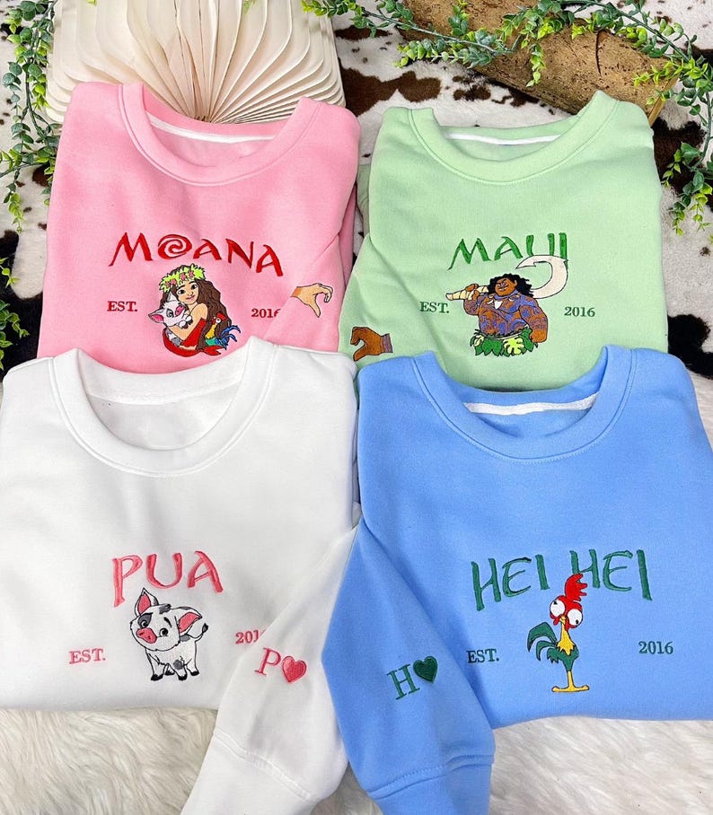 May include: Four crewneck sweatshirts in pink, light green, white, and blue. Each features embroidered character art and text: Moana, Maui, Pua, or Hei Hei. The text includes names, EST. 2016, and small sleeve details like a heart or hand icon.