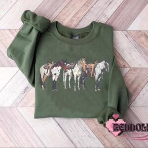 May include: A green sweatshirt with a graphic of four horses with saddles. The horses are white, brown, and black.