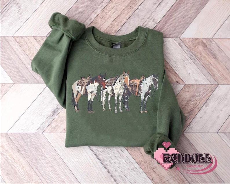 May include: A green sweatshirt with a graphic of four horses with saddles. The horses are white, brown, and black.