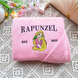 Może przedstawiać: Złożony r&oacute;żowy koc z haftowanym wzorem Roszpunki z długimi blond włosami i zielonym ptakiem. Słowo "RAPUNZEL" jest wygięte nad obrazem, z "EST. 2010" poniżej.