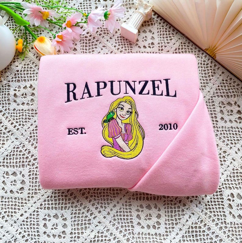 Może przedstawiać: Złożony r&oacute;żowy koc z haftowanym wzorem Roszpunki z długimi blond włosami i zielonym ptakiem. Słowo "RAPUNZEL" jest wygięte nad obrazem, z "EST. 2010" poniżej.