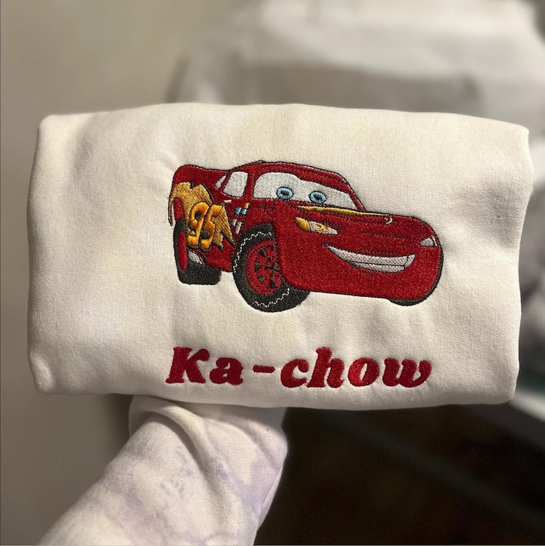 May include: A white sweatshirt featuring an embroidered red race car with the number 95 in yellow on the side. Below the car, the text Ka-chow is embroidered in red thread. The design includes blue eyes, a silver mouth, and black tires.