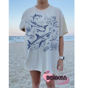 K&ouml;nnte beinhalten: Ein wei&szlig;es T-Shirt mit einem blauen Grafikdruck verschiedener Meeresbewohner, darunter Haie, Quallen und ein Tintenfisch. Die Grafik ist mittig auf dem Shirt platziert und bedeckt den gr&ouml;&szlig;ten Teil der Vorderseite.