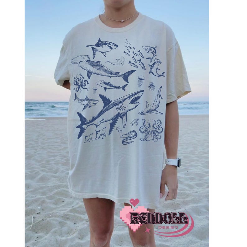 K&ouml;nnte beinhalten: Ein wei&szlig;es T-Shirt mit einem blauen Grafikdruck verschiedener Meeresbewohner, darunter Haie, Quallen und ein Tintenfisch. Die Grafik ist mittig auf dem Shirt platziert und bedeckt den gr&ouml;&szlig;ten Teil der Vorderseite.