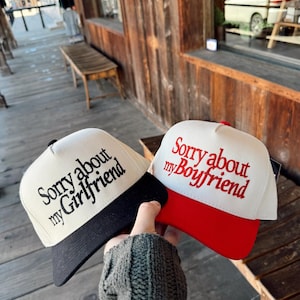 May include: Two baseball caps with the text "Sorry about my Girlfriend" and "Sorry about my Boyfriend". One cap has a black brim, the other a red brim. Both caps have a white crown.