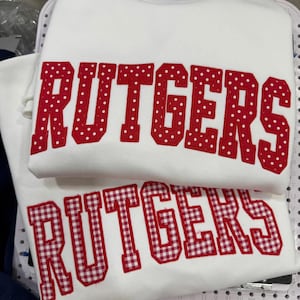 May include: A white sweatshirt featuring the word RUTGERS in large, red, polka-dot fabric letters. Below it, a second white garment displays the word RUTGERS in red and white gingham fabric letters, both applied with a visible stitched border.